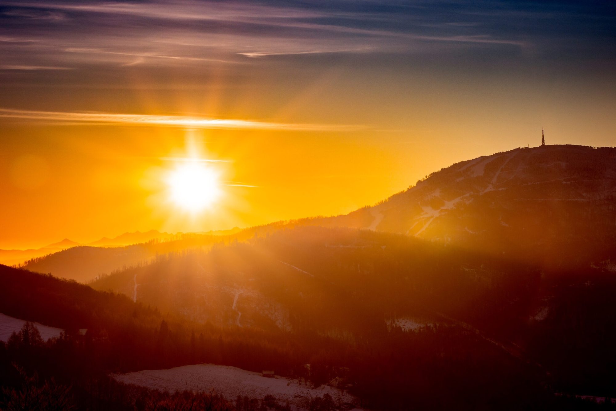 The sun coming up behind a forested mountain ridge, mist lying along the valley floor below.