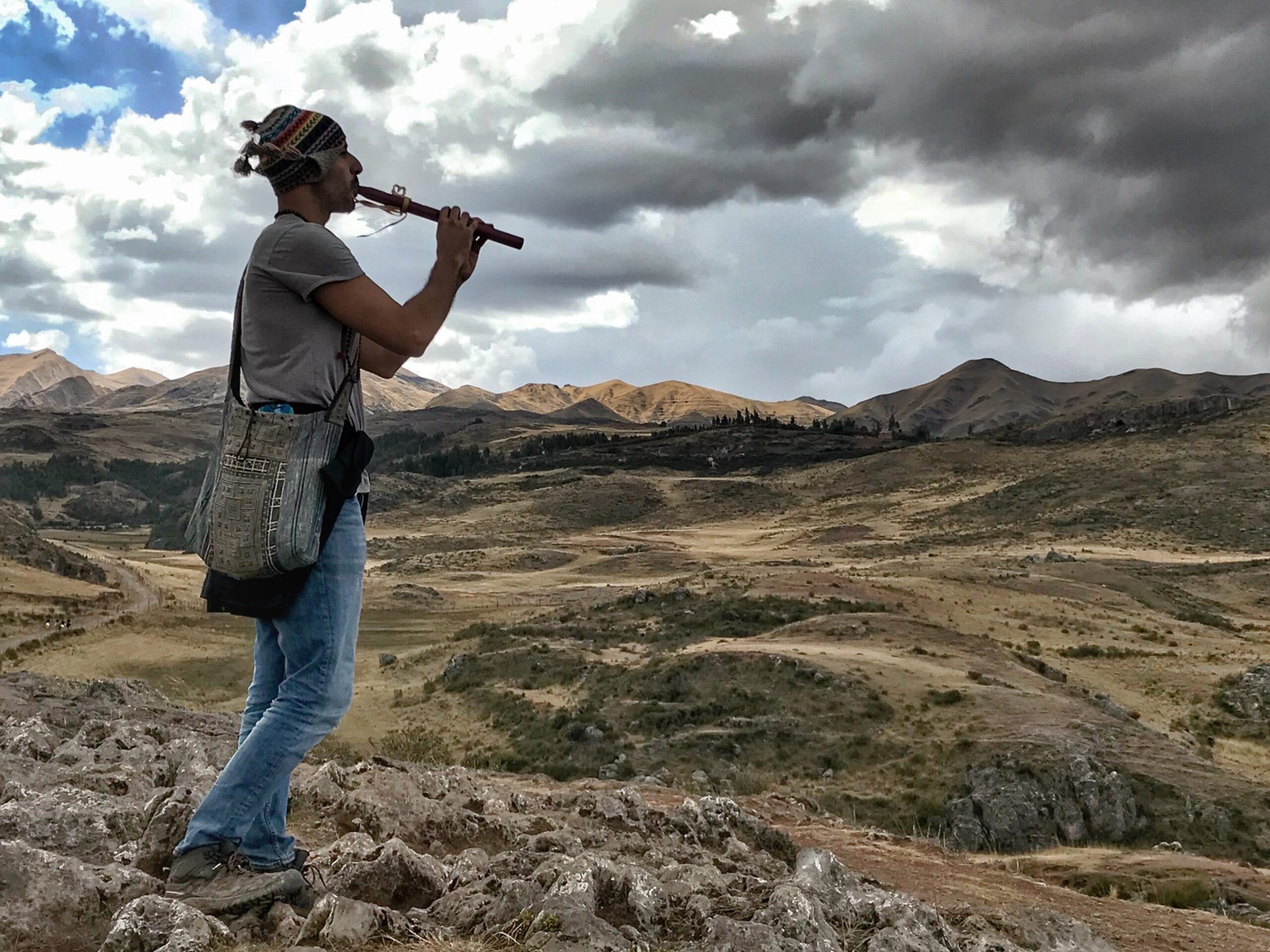 Hamid Jabbar playing a wooden flute on a rocky ridge, a woven bag over his shoulder, golden grassland and bare mountains behind him under a clouded sky.