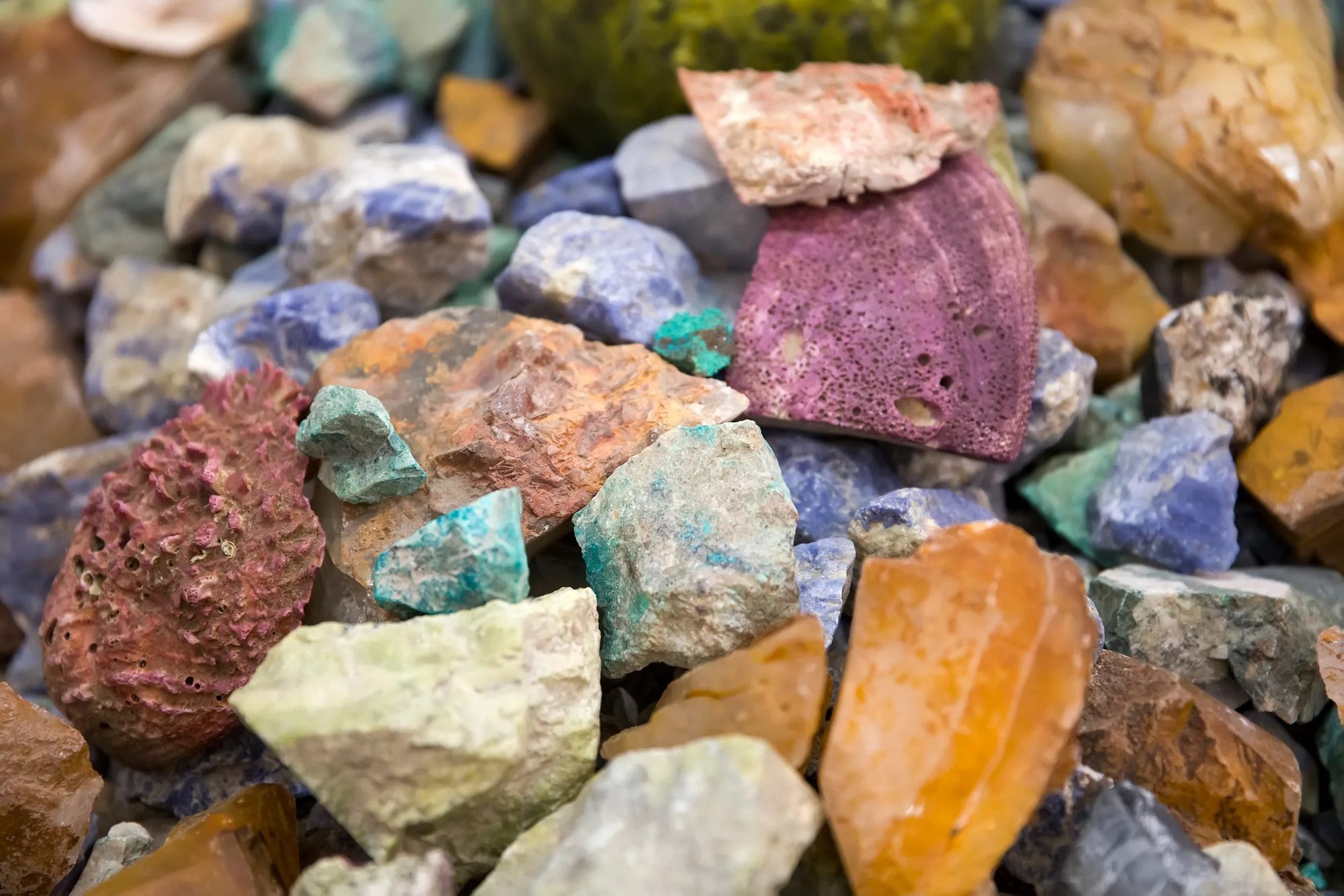 A heap of raw uncut mineral stones, pink, turquoise, amber and blue, broken faces catching the light.