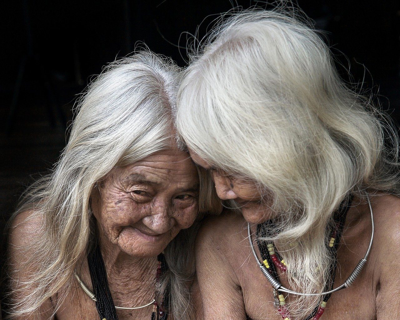 Two elderly women with long white hair, leaning in until their foreheads touch, both laughing.