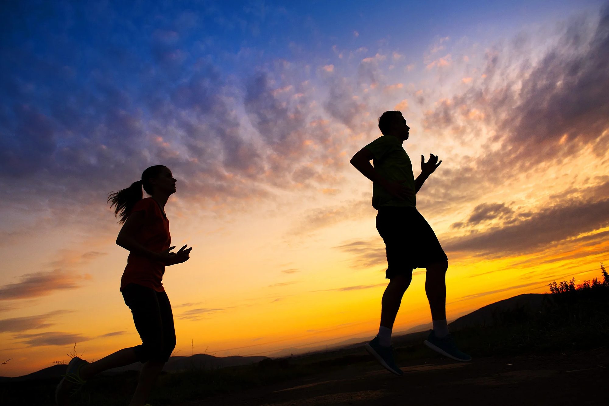 Two people running uphill in silhouette against an orange and blue sky at the end of the day.