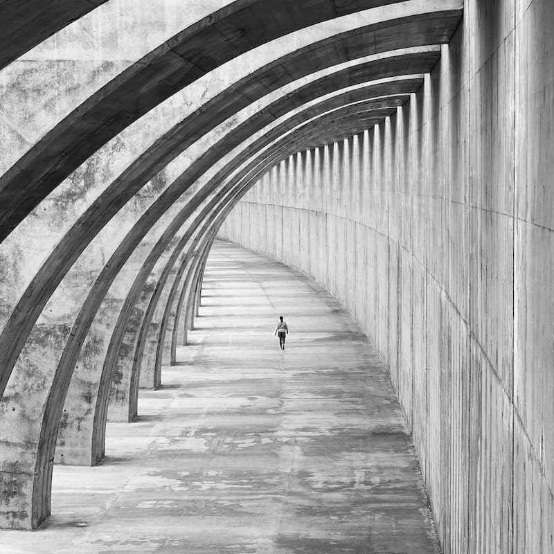 A black and white photograph of a lone figure walking down a long, curving concrete passage lit through a row of arches.