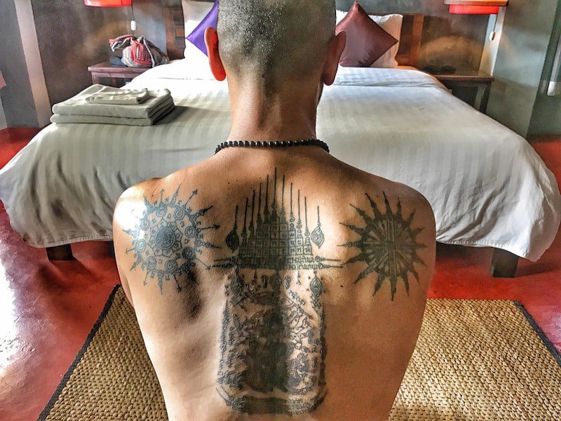A man with a shaved head sits with his bare back to the camera, covered in Thai Sak Yant tattoos, a tall yantra of spires down his spine and a round sunburst on each shoulder, facing a made bed.