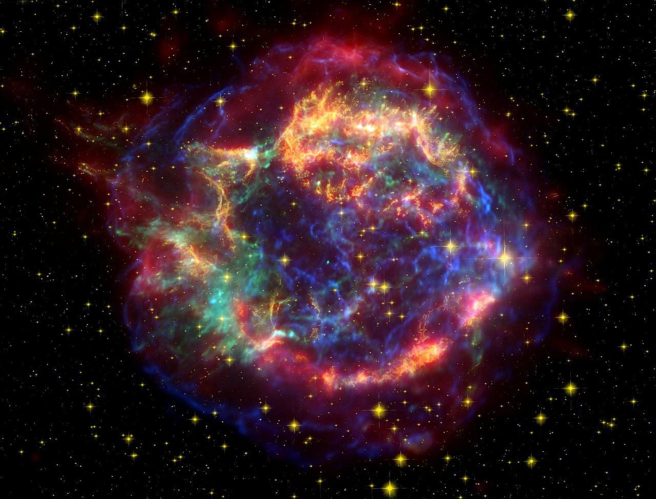A glowing ring of red, orange, green and blue gas left by an exploded star, on a black sky full of yellow stars.