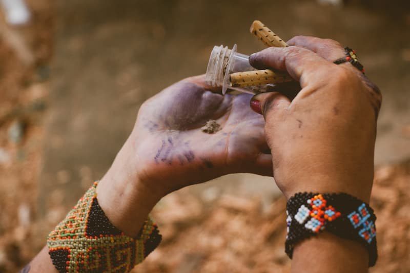 Two hands in beaded bracelets, one holding a small vial and a carved wooden pipe, the other a pinch of brown rapé powder in a palm stained purple.