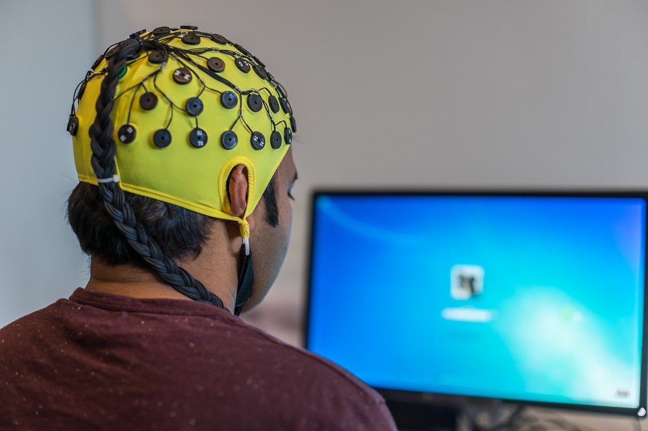 A man seen from behind, his hair in a long braid, wears a yellow EEG cap covered in electrodes and wires as he faces a computer screen.