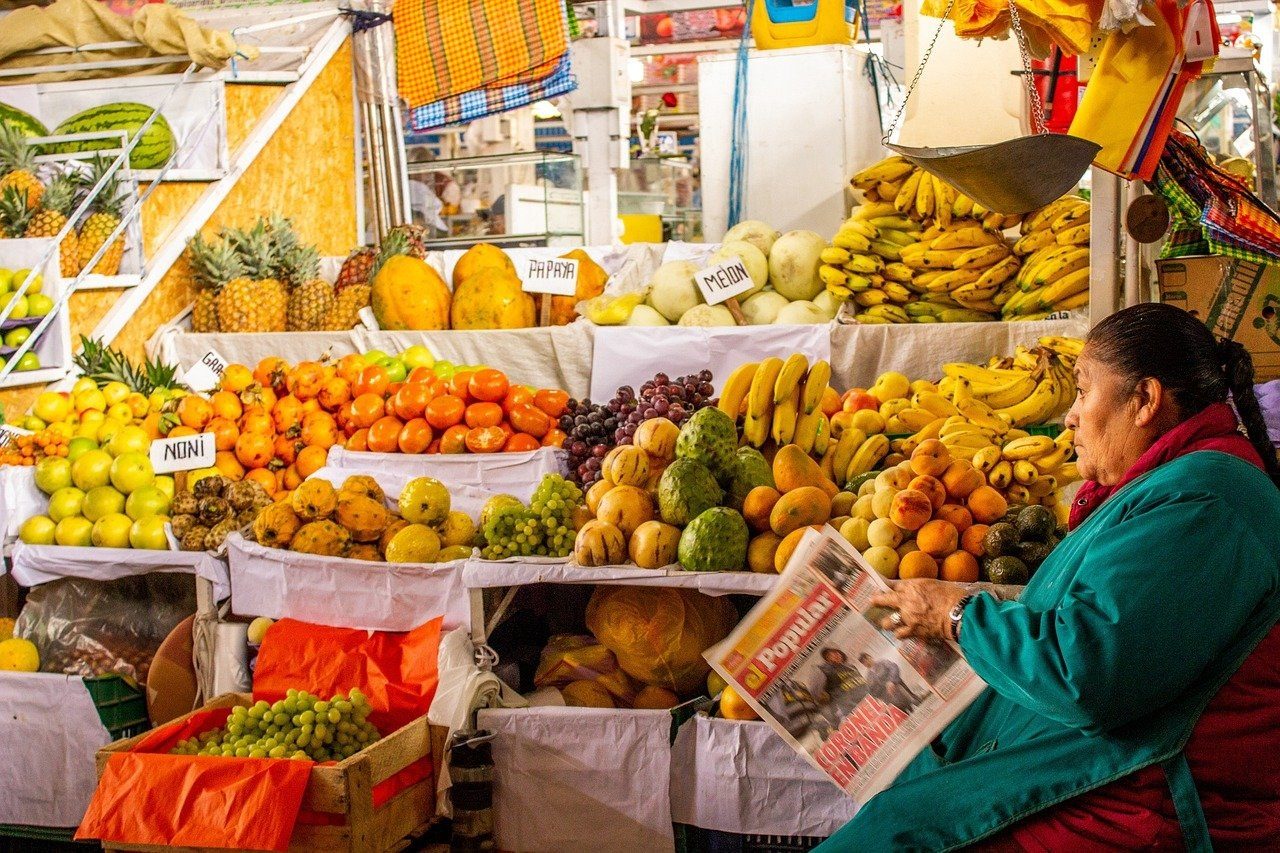 A market stall piled with pineapples, papayas, melons, bananas, oranges and grapes, with a woman in a teal jacket reading a newspaper at its right edge.