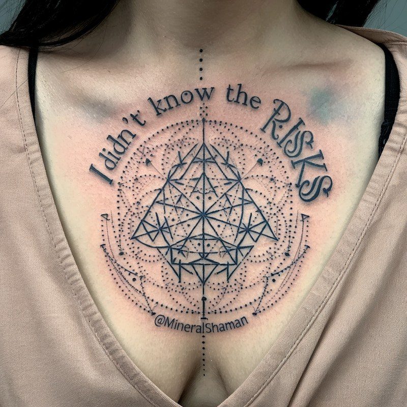 A black ink tattoo on a woman’s upper chest, a triangle of fine lines inside rings of dots, with the words “I didn’t know the risks” arched above it and “@MineralShaman” below.
