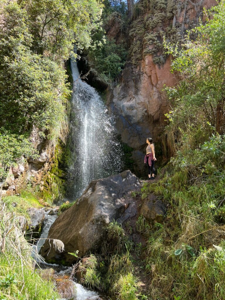 A woman standing on a boulder beside a tall waterfall in a green gorge, looking up at the falling water.