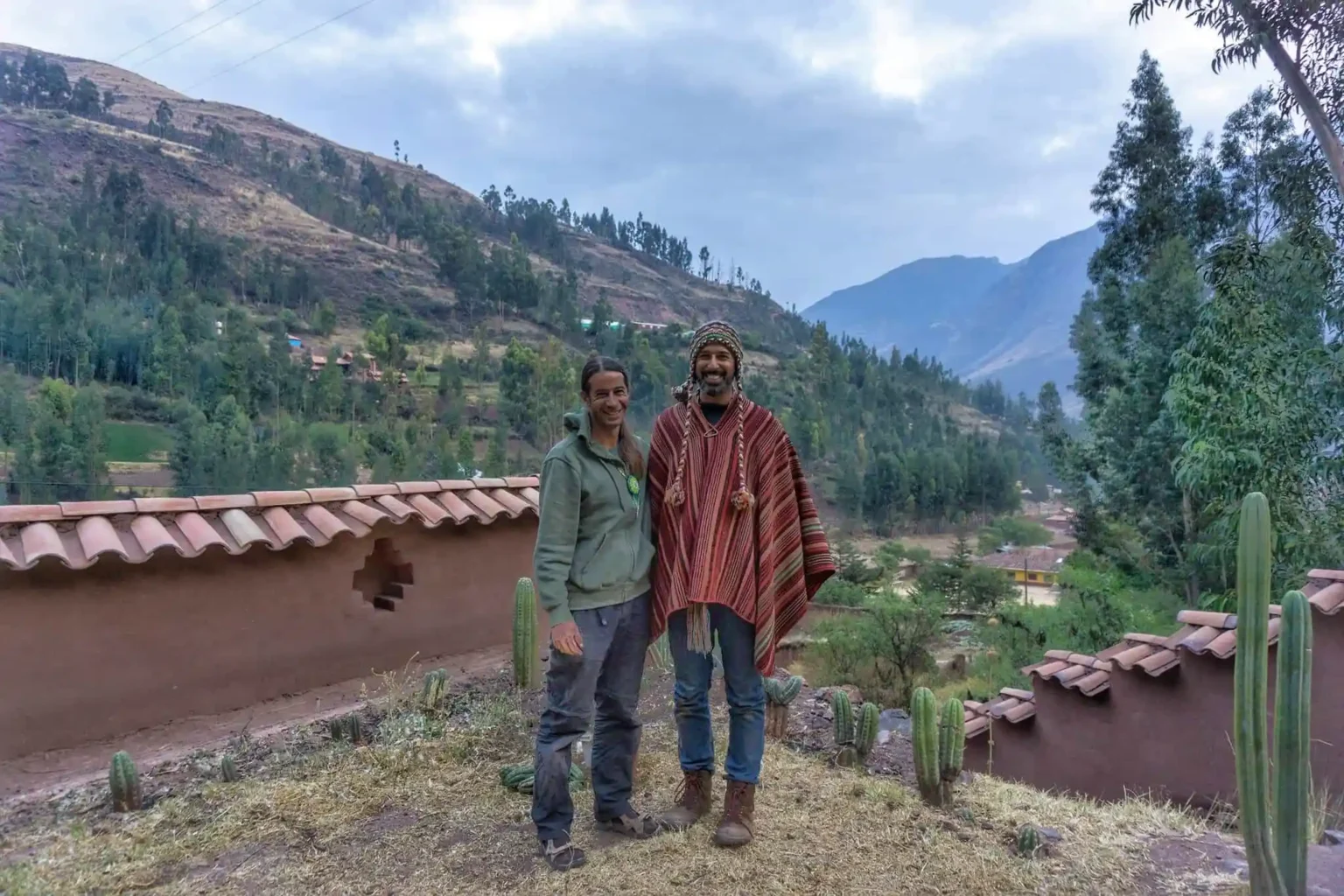 Two men standing at a low adobe wall above the valley, one in a striped poncho and knitted chullo, young cacti planted along the wall.