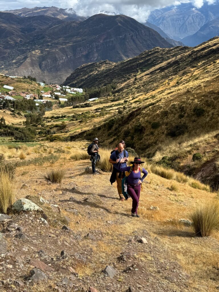 Three people climbing a grassy ridge high above a valley village, one turning back toward the camera with a hand raised.