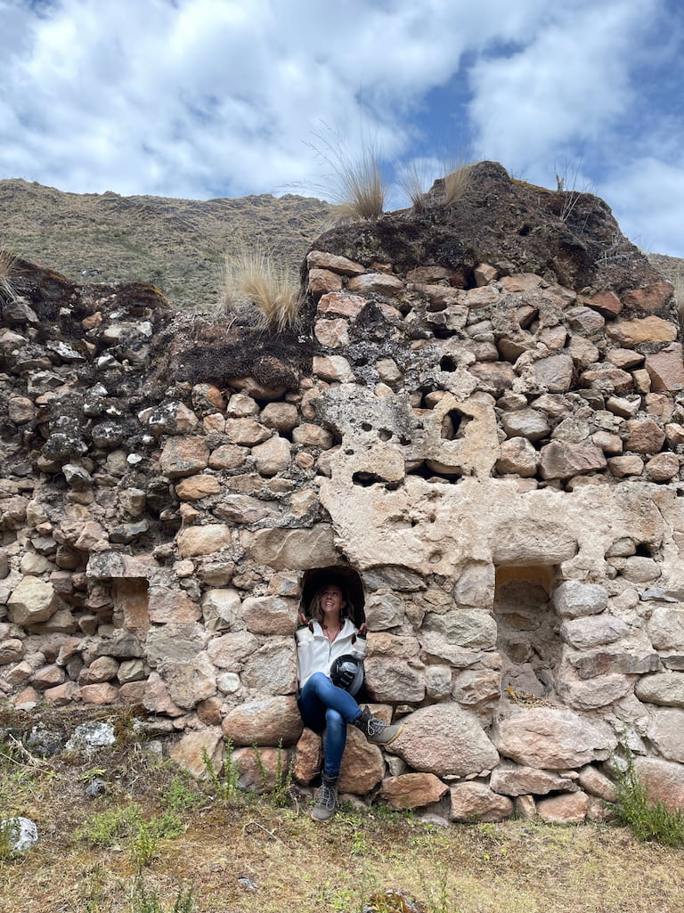 A woman sitting in a doorway of a dry stone ruin on a hillside, the wall rising above her with grass growing out of its top.