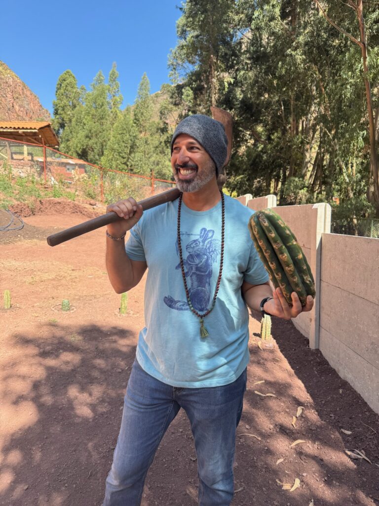 Hamid Jabbar laughing with a pickaxe over one shoulder and cut lengths of San Pedro cactus under the other arm, at the planting ground.