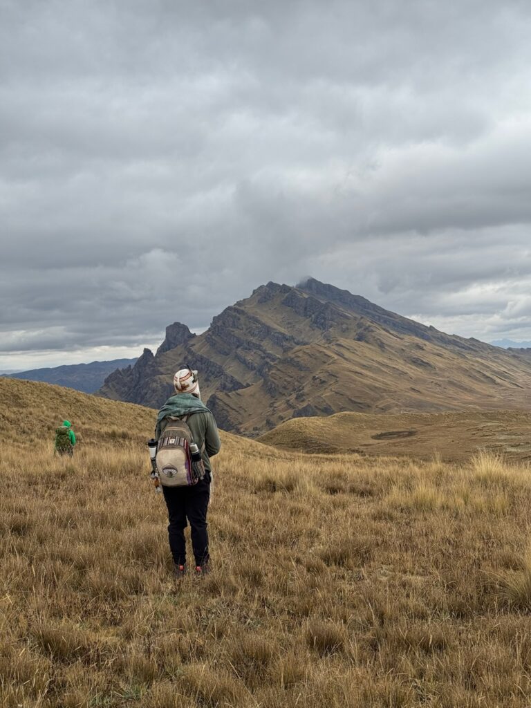 Two people crossing high golden grassland toward a steep rocky peak, the nearer one carrying a pack and a thermos.
