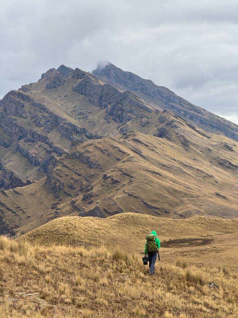 A person in a green hooded jacket walking alone across high grassland toward a steep rock face.