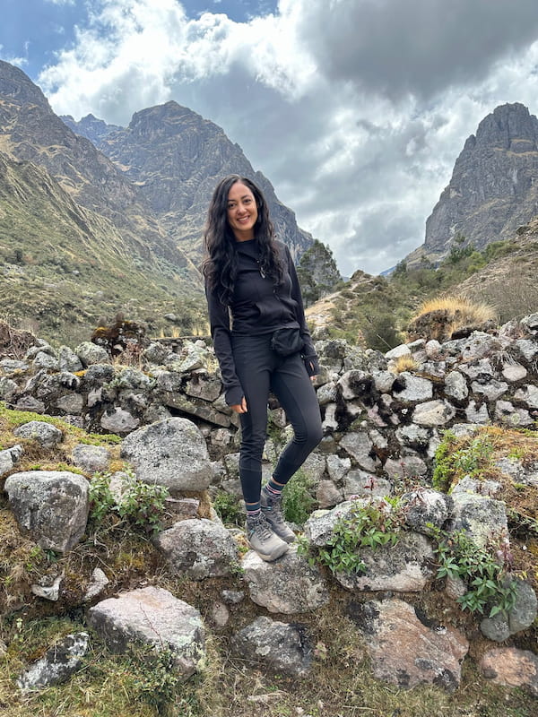 A woman standing on an old stone wall in a steep Andean valley, rock peaks rising on both sides behind her.
