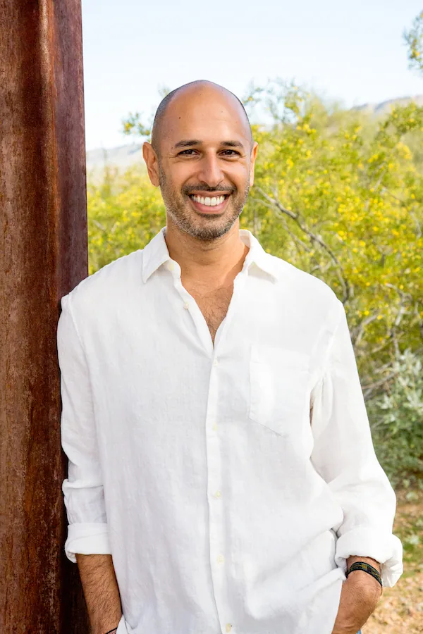 Hamid Jabbar in a white linen shirt, leaning against a weathered post with desert scrub and yellow blossom behind him.