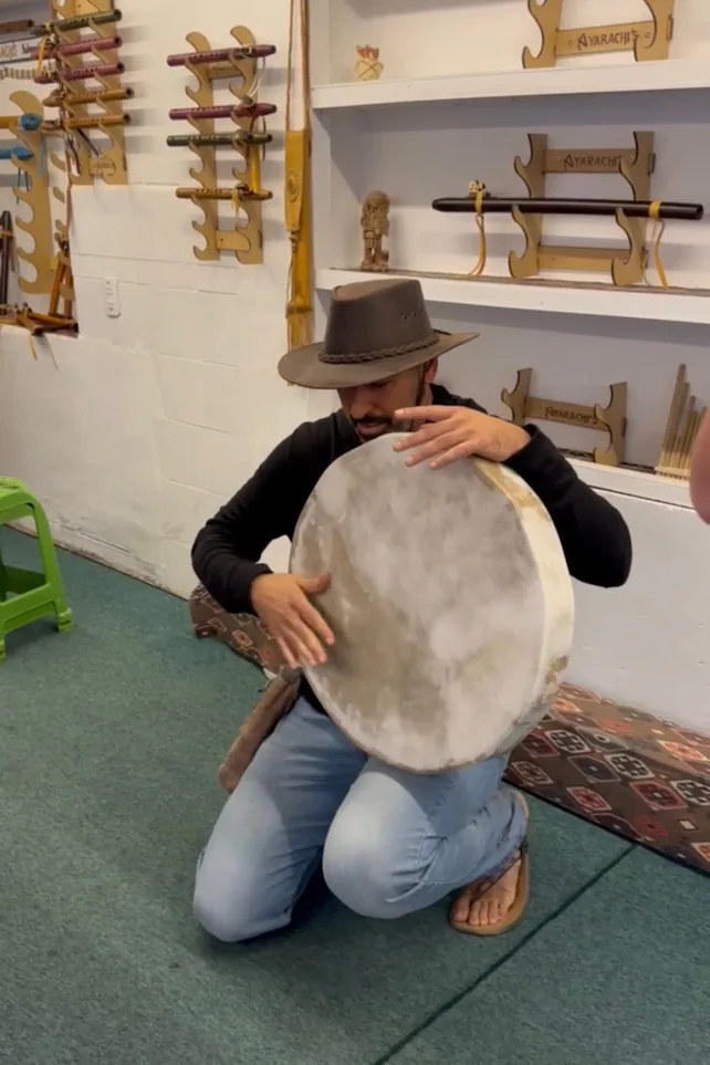 A man kneeling to play a large frame drum in a workshop hung with Andean flutes and other instruments.