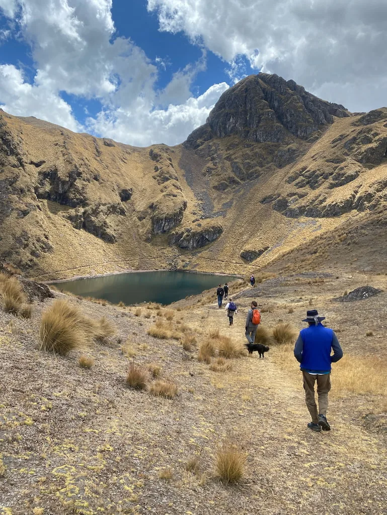 Retreat guests walking down a grass slope toward a small dark lake ringed by high Andean ridges, a dog trotting alongside them.