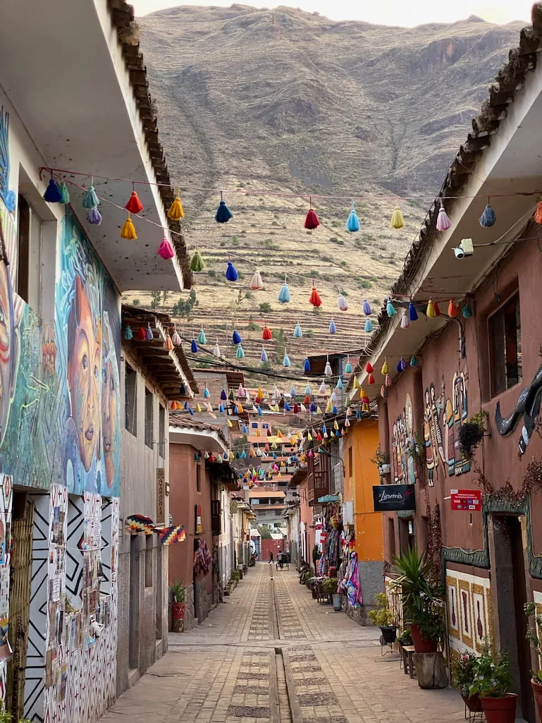 A narrow street in Pisac hung with rows of coloured tassels, painted shopfronts on both sides and terraced mountainside beyond.