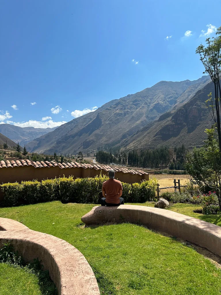 A man in an orange shirt sitting cross legged on a curved earthen bench, facing a green lawn, tiled roofs and the mountains across the Sacred Valley.