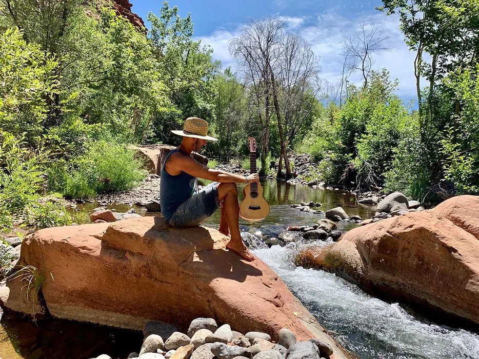 Hamid Jabbar sitting on red rock beside a running creek with a small guitar on his knee, wearing a straw hat.